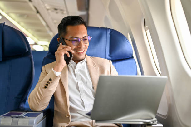 Professional businessman in beige suit working on laptop and talking on phone while seated in an airplane's business class cabin.